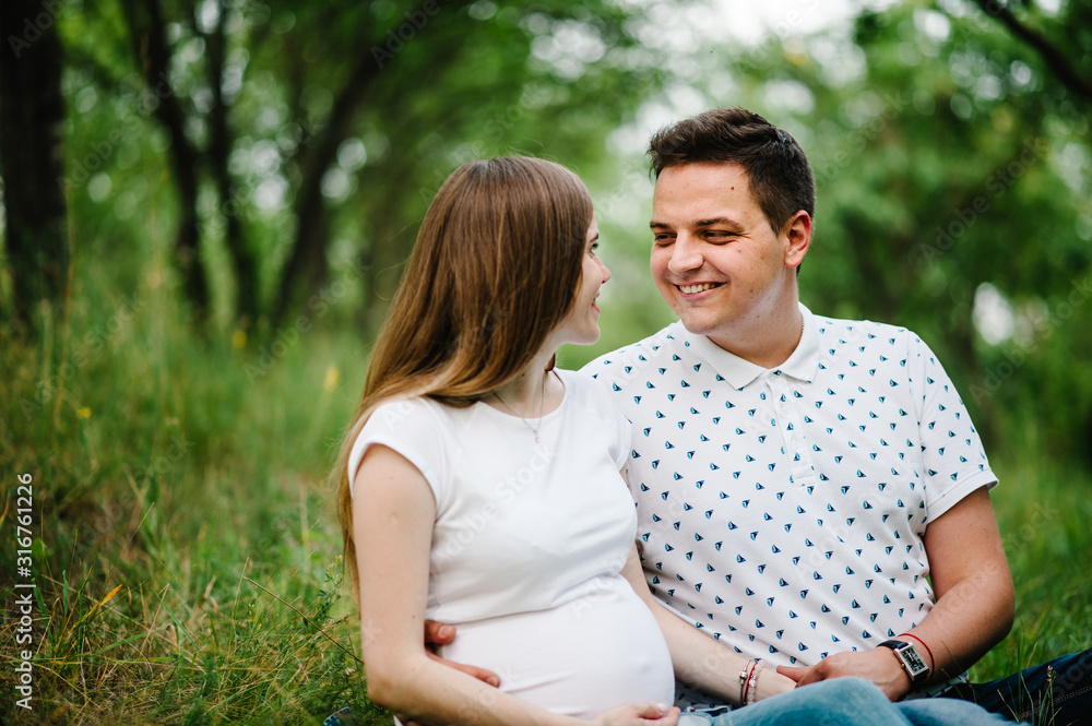 Pregnant girl and her husband are happy to hug, hold hands on stomach, sitting on the grass in the outdoor in the garden background. Close up. upper half. Look at each other.