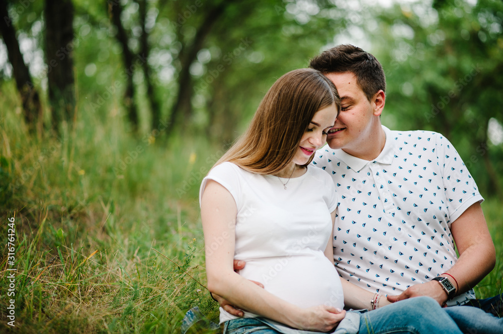 Pregnant girl and her husband are happy to hug, hold hands on stomach, sitting on the grass in the outdoor in the garden background. Close up. upper half. Looking down Pregnant girl and her husband are happy to hug, hold hands on stomach, sitting on the grass in the outdoor in the garden background. Close up. upper half. Looking down