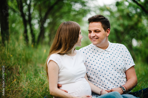Pregnant girl and her husband are happy to hug, hold hands on stomach, sitting on the grass in the outdoor in the garden background. Close up. upper half. Look at each other.