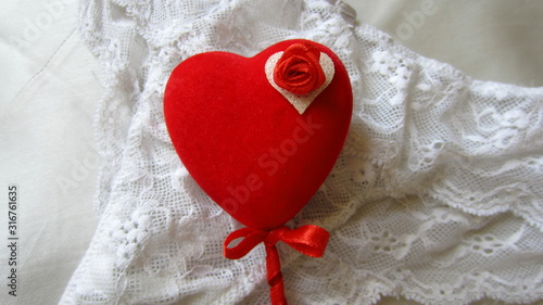 A red heart on a stick with a small red bow, decorated with a small red rose, lying on a white lace underwear. Saint Valentine's Day sexy gift concept.