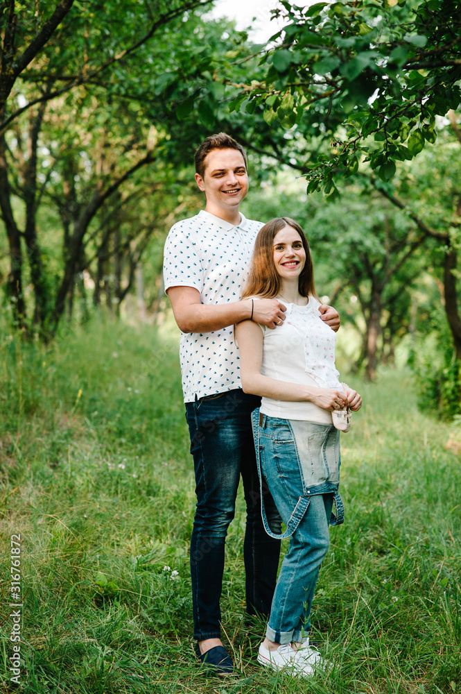 Pregnant girl and husband hold hands hug, to put baby shoes and clothes on stomach, waiting baby. stand in the outdoor in the garden background. Close up. full length. looking at camera Pregnant girl and husband hold hands hug, to put baby shoes and clothes on stomach, waiting baby. stand in the outdoor in the garden background. Close up. full length. looking at camera