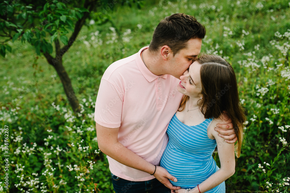 Pregnant girl and her husband kiss are happy to hug, hold hands on stomach, stand in the outdoor in the garden background with trees. Close up. upper half. Pregnant girl and her husband kiss are happy to hug, hold hands on stomach, stand in the outdoor in the garden background with trees. Close up. upper half.