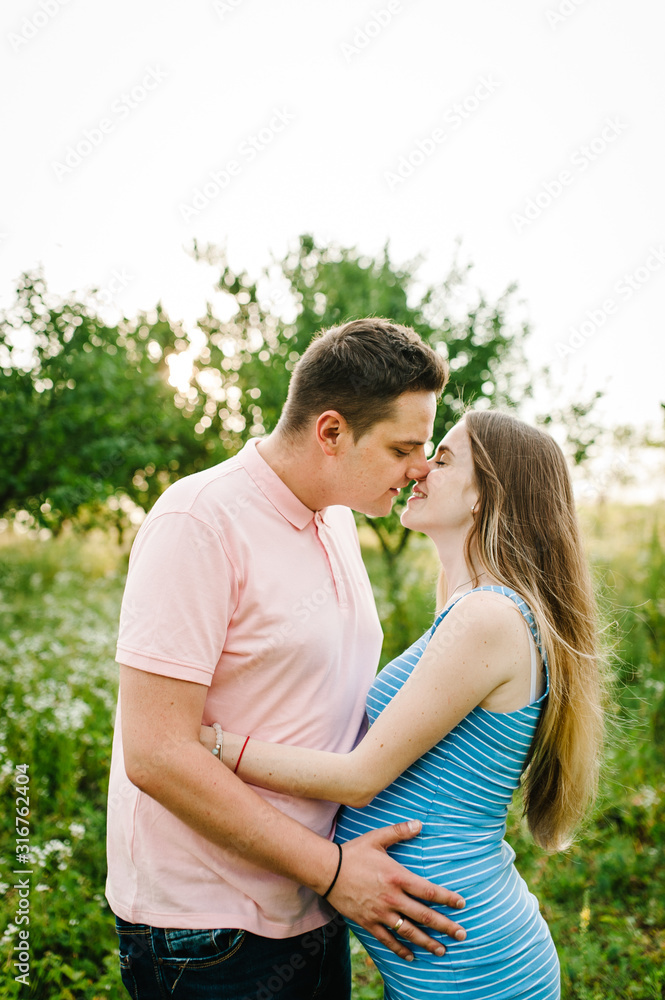Pregnant girl and her husband to hug and kiss, hold hands round stomach, stand on grass in the outdoor in the garden background with trees. Close up. length upper half. Closed eyes