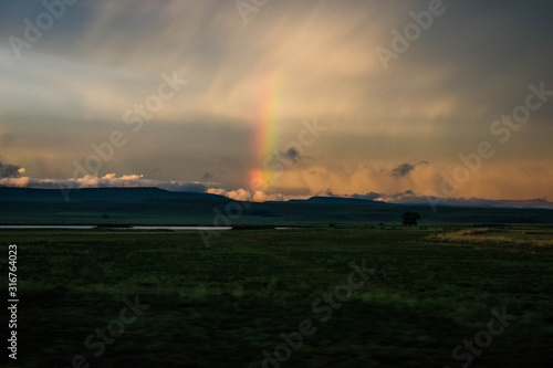 grasslands view of rainbow in between mountains and clouds