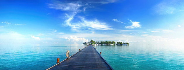 Beautiful tropical landscape background, concept for summer travel and vacation. Wooden pier to an island in ocean against blue sky with white clouds, panoramic view.