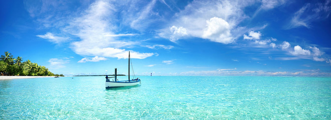 Boat in turquoise ocean water against blue sky with white clouds and tropical island. Natural landscape for summer vacation, panoramic view.