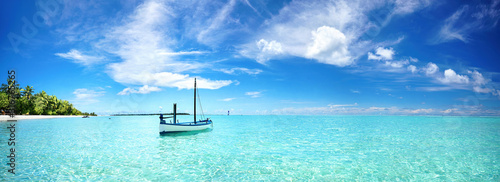 Boat in turquoise ocean water against blue sky with white clouds and tropical island. Natural landscape for summer vacation, panoramic view. © Laura Pashkevich