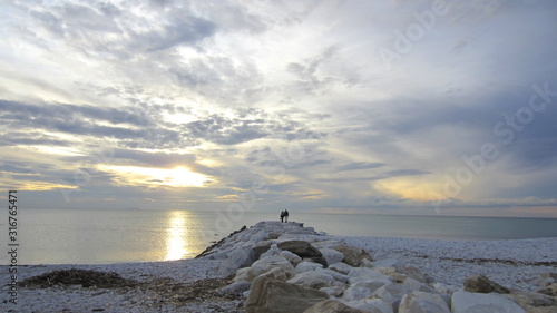 The couple watches the sunset over the sea, standing on a rocky waterfront. Cloudy pastel sky and romantic atmosphere