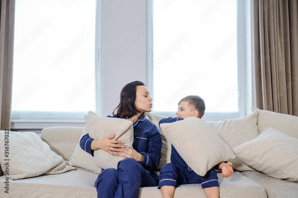 Happy young mother lying on bed and playing/having fun with pillows with her son. Both a wearing the same blue pyjamas, happy time on bed