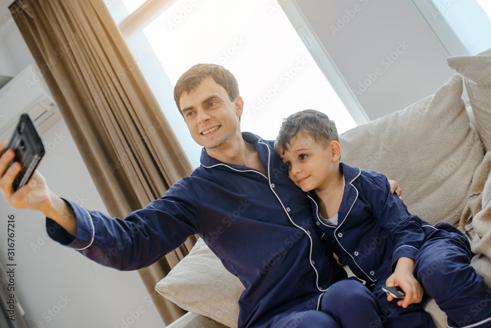 Fototapeta premium Happy young father making selfie with camera smartphone and playing with his son. Both a wearing the same blue pyjamas