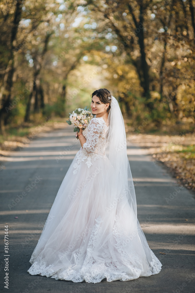 Beautiful and smiling bride in a white dress with a bouquet walks along the road in the forest with yellow trees. Autumn wedding portrait. Photography, concept.