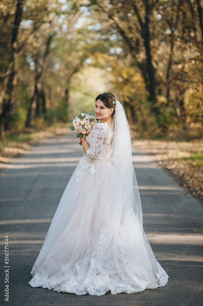 Beautiful and smiling bride in a white dress with a bouquet walks along the road in the forest with yellow trees. Autumn wedding portrait. Photography, concept. Beautiful and smiling bride in a white dress with a bouquet walks along the road in the forest with yellow trees. Autumn wedding portrait. Photography, concept.