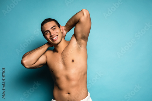 Fitness and sports. Beauty portrait of half naked handsome smiling young man with beautiful torso dressed in towel showing his biceps looking at camera isolated over blue background