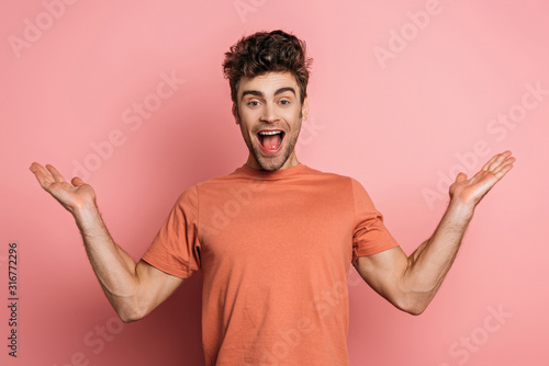 excited young man laughing at camera while standing with open arms on pink background