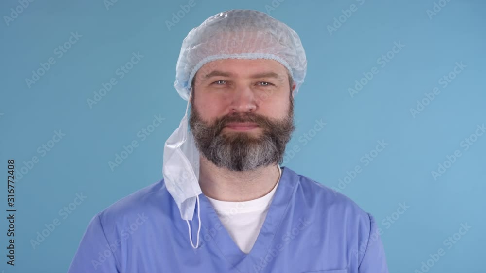 Dolly-out portrait shot of middle-aged male doctor wearing scrubs, bouffant cap and gloves standing isolated on blue background and taking off procedure mask, then smiling for camera Dolly-out portrait shot of middle-aged male doctor wearing scrubs, bouffant cap and gloves standing isolated on blue background and taking off procedure mask, then smiling for camera