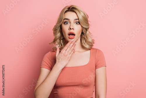 shocked girl looking at camera while holding hand near open mouth on pink background