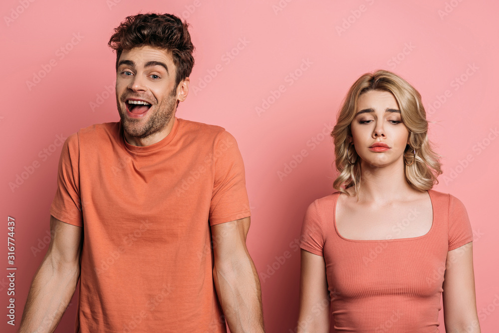 cheerful man looking at camera while standing near offended girlfriend on pink background cheerful man looking at camera while standing near offended girlfriend on pink background