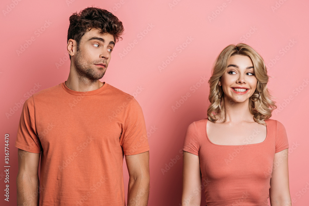 thoughtful man looking at cheerful girlfriend on pink background thoughtful man looking at cheerful girlfriend on pink background
