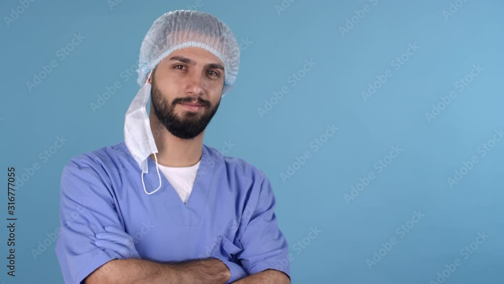 Dolly-in portrait shot of bearded male doctor in uniform, procedure mask and bouffant cap standing isolated on blue background and crossing his arms while staring at camera Dolly-in portrait shot of bearded male doctor in uniform, procedure mask and bouffant cap standing isolated on blue background and crossing his arms while staring at camera