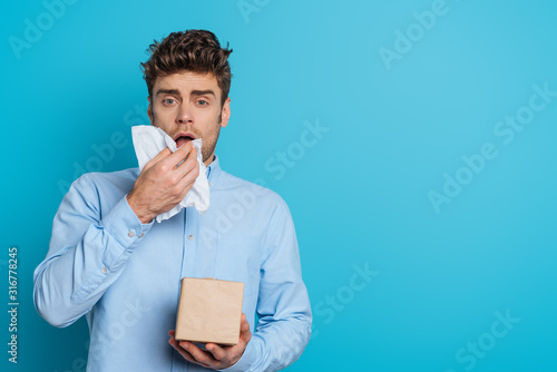 sick man sneezing in paper napkin while looking at camera on blue background photo