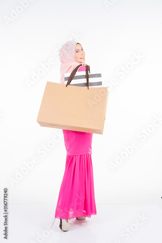 A beautiful and excited Muslim female model in a traditional pink modern kurung carrying shopping bags isolated on white background. Eidul fitri fashion and festive shopping concept © HEMINXYLAN