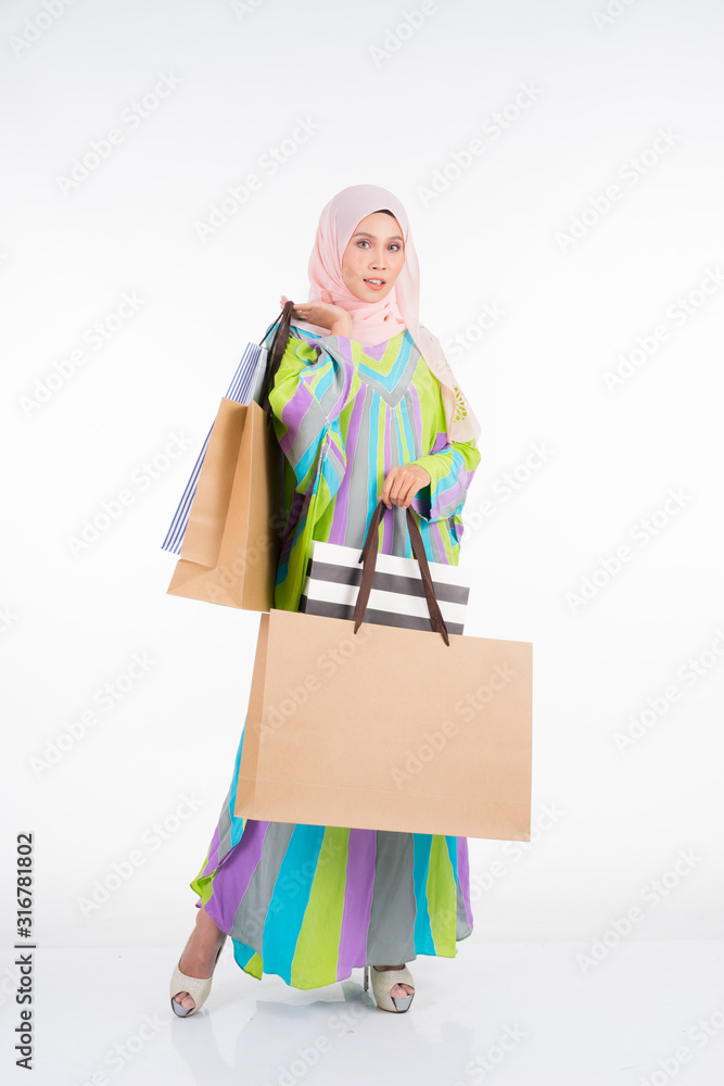 Naklejka premium A beautiful and happy Muslim female model in a traditional kaftan/caftan dress carrying shopping bags isolated on white background. Eidul fitri fashion and festive preparation shopping concept