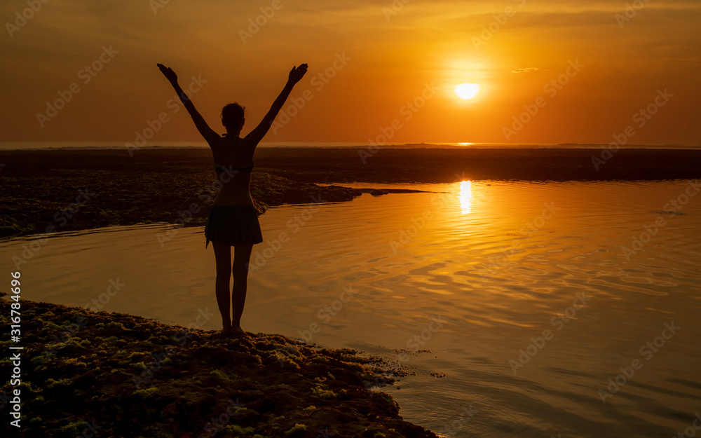 Excited young woman raising arms at the beach in front of the ocean. View from back. Sunset golden hour at the beach. Bali, Indonesia.