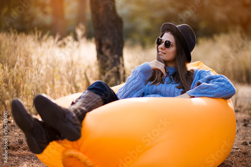 Autumn lifestyle portrait of pretty girl in stylish sunglasses and black hat sitting on orange inflatable sofa at forest sunset. Relaxing and enjoying life on air bed photo