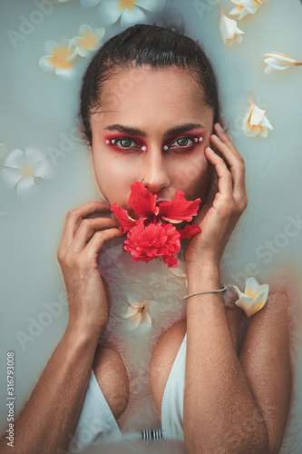 Fashion portrait of beautiful woman with red flower in her mouth and white flowers in a bath with milk