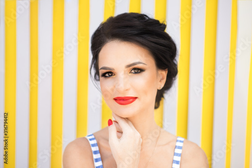 Horizontal portrait of beautiful young brunette smiling woman with red lipstick and red nails in striped blue dress on striped yellow background in summertime. Hot summer girl with sexy smile.