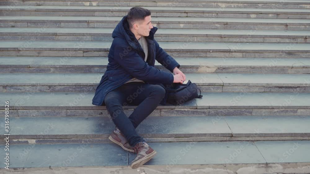 A young man is sitting on the stairs and using his smartphone. Then he is going out the focus. Copy space. Action. Animation. 4K. A young man is sitting on the stairs and using his smartphone. Then he is going out the focus. Copy space. Action. Animation. 4K.