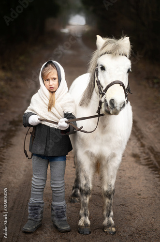 A little village girl in modest clothes walks in the winter with her little white pony in the forest. Original image. Country life.