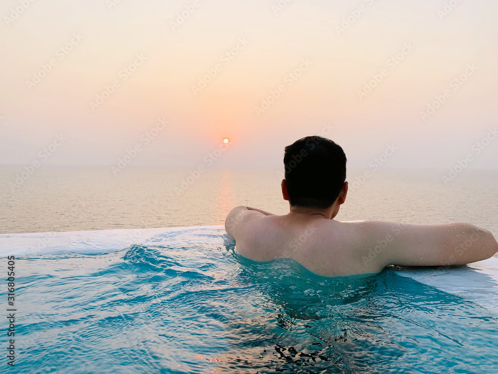 Young healthy man soaking and swimming in an infinity pool with beautiful and peaceful ocean and sunset view during his holiday. Solo travel for weekend getaway and lifestyle concept
