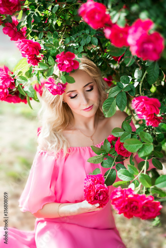a blond girl surrounded by roses with her eyes closed inhales the scent of beautiful flowers. Bright makeup and a pink dress on her