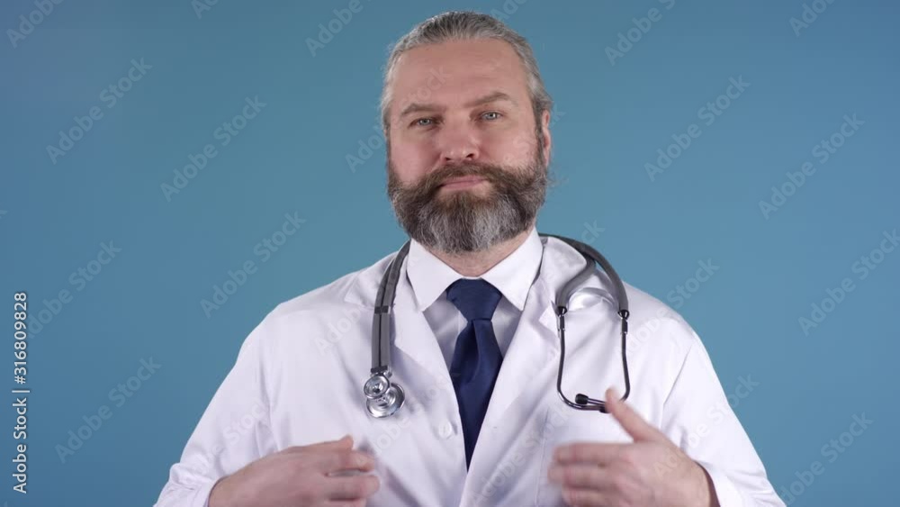 Dolly-in portrait of bearded middle-aged male doctor in white coat standing isolated on blue background and placing stethoscope around his neck while looking at camera Dolly-in portrait of bearded middle-aged male doctor in white coat standing isolated on blue background and placing stethoscope around his neck while looking at camera