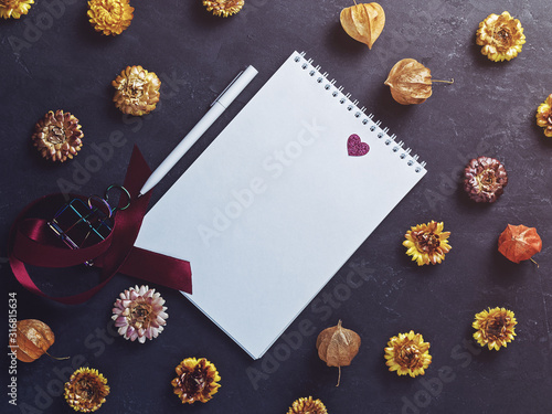 Background for Valentine's Day. A notebook with blank sheets is on a dark background. Nearby are dried flowers and a white pen. Flat lay, copy space.
