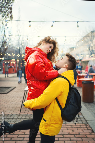 young romantic couple in love hugging, wearning in bright yellow and red down jackets. Handsome man carrying young attractive woman while walking on the street. spending time together, having a date photo