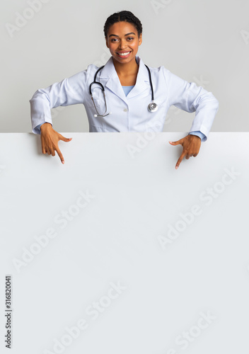 Smiling afro female intern doctor pointing at blank advertisement board photo