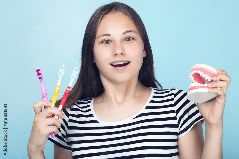 Young girl with dental braces holding teeth model and toothbrushes on blue background Young girl with dental braces holding teeth model and toothbrushes on blue background