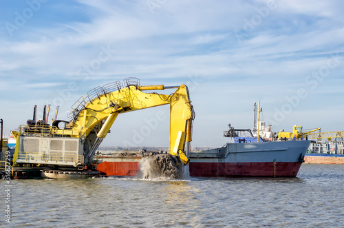 Deepening the water area of the port. A floating excavator loads soil onto a barge.