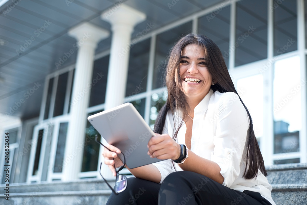 rub a beautiful young business lady, smiling, holding a tablet. Sits on the steps of an office building