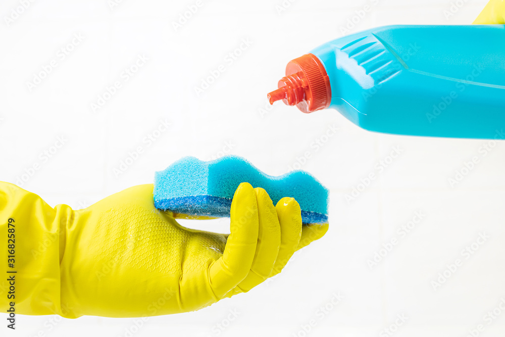 Female hand in a yellow latex glove with a blue sponge. In the other hand is a bath cleaner. Background - white tile. The concept of cleaning the bathroom, cleanliness. Copyspace.