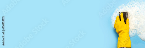 Hand in a yellow rubber glove holds a cleaning sponge and wipes a soapy foam on a blue background. Cleaning concept, cleaning service. Banner. Flat lay, top view
