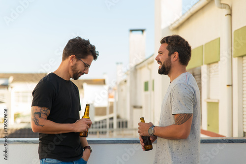 Two handsome young men talking while drinking beer. Smiling friends communicating on balcony. Leisure, friendship concept