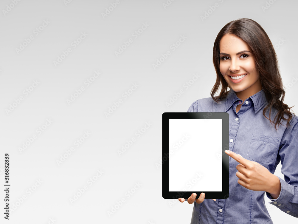 Happy smiling attractive businesswoman showing blank tablet pc monitor, with copy space area for some text, advertising or slogan, over grey background - obrazy, fototapety, plakaty 
