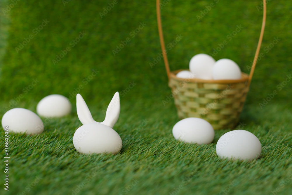 White chicken eggs on the green grass, behind which the ears of the Easter rabbit are hidden. Wicker basket. Symbols for the celebration of religious holiday among Christians and Catholics. White chicken eggs on the green grass, behind which the ears of the Easter rabbit are hidden. Wicker basket. Symbols for the celebration of religious holiday among Christians and Catholics.