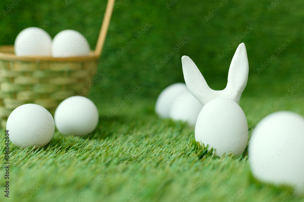 White chicken eggs on the green grass, behind which the ears of the Easter rabbit are hidden. Wicker basket. Symbols for the celebration of religious holiday among Christians and Catholics. White chicken eggs on the green grass, behind which the ears of the Easter rabbit are hidden. Wicker basket. Symbols for the celebration of religious holiday among Christians and Catholics.