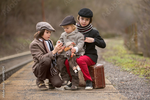 Adorable boy on a railway station, waiting for the train with suitcase and beautiful vintage doll...