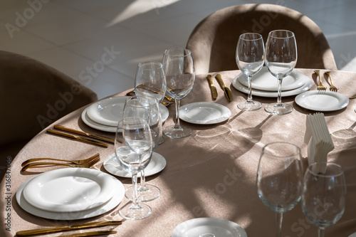Chic and elegant, gold-plated cutlery and white plates, table setting with empty plates. Glasses in the light from the window.