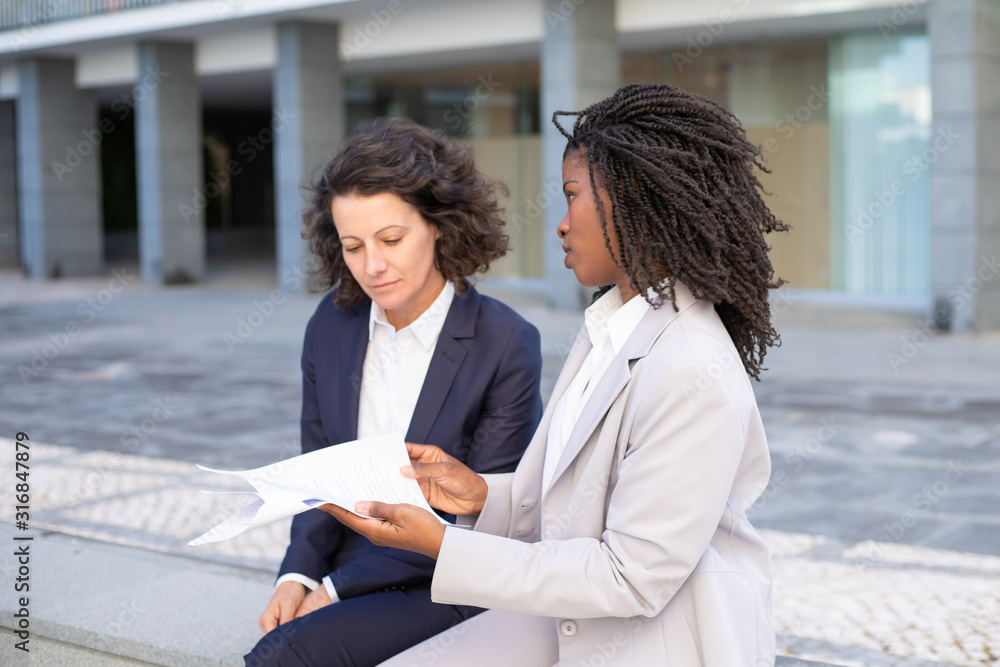 Serious businesswomen discussing contract. Professional multiethnic businesswomen sitting on street and working with documents. Paperwork concept Serious businesswomen discussing contract. Professional multiethnic businesswomen sitting on street and working with documents. Paperwork concept
